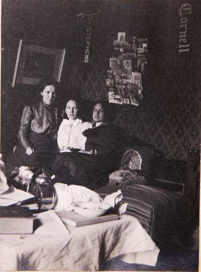 Three women in what looks like a college students room from 1910. Banners from nearby schools are on the wall. A bulletin board full of photos is on the wall. A pillow with the univeristy logo is in the photos. And there are piles of books and papers, etc