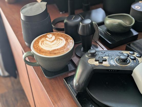 A coffee with latte art sitting next to a Legend of Zelda: Tears of the Kingdom special edition switch pro controller. They're on a walnut coffee bar surrounded by matte black and walnut coffee gear.