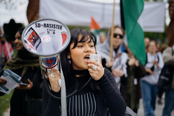 A student protester on megaphone, we can see a group of protesters behind her, marching with Palestine's flag. The megaphone has many stikers, "SD=RASISTER", "General Strike".