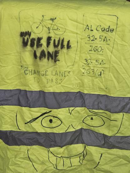 A bright yellow vest with 2 silver reflective stripes. In black magic marker are the following: at the bottom are eyes and a smiling mouth with teeth. On top are 3 panels. The 1st panel has a drawing of a bicycle. Under it says "May use full lane". The 2nd panel say "Change lanes to pass". The 3rd panel says "AL Code 32-5a 260 32-5a 263(a)