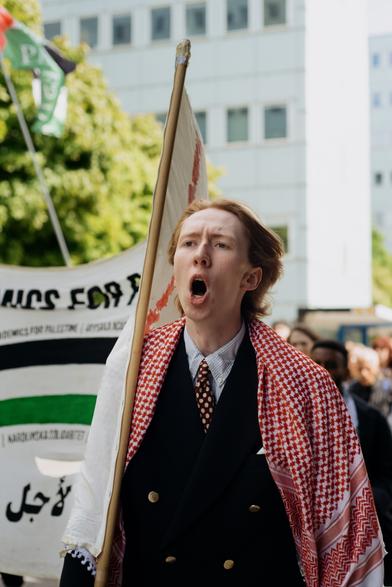 A protester wearing a red keffiyeh holding one side of the banner and shouting the slogans while marching.