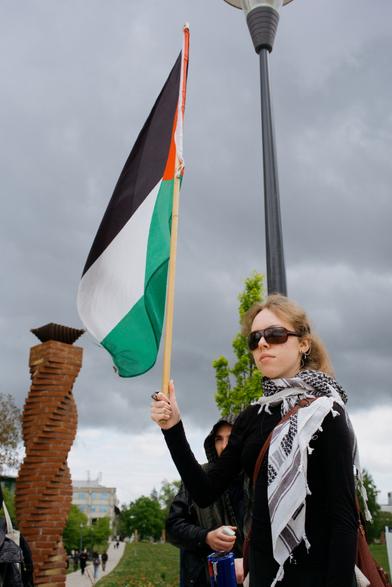 A protester is holding a large Palestine flag while wearing a kaffiyeh, we can see dark cloud above in the sky.