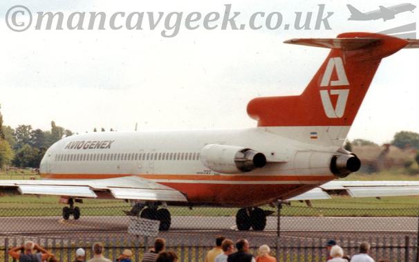 Side view of a 3 engined jet airliner with the engines mounted on the rear of the fuselage, taxiing from right to left as it turns from a taxiway on to a runway.
The plane is mostly white, with an orange stripe running along the lower body, and a red belly.
There are small, black "Aviogenex" titles on the upper forward fuselage.
The tail is red, with a stylised white letter "A", with a reflection below.
A black chain-link fence stretches across the bottom of the frame, with people standing around, some looking at the plane.
Green grass on the far side of the runway leads off to trees in the distance, under grey skies.