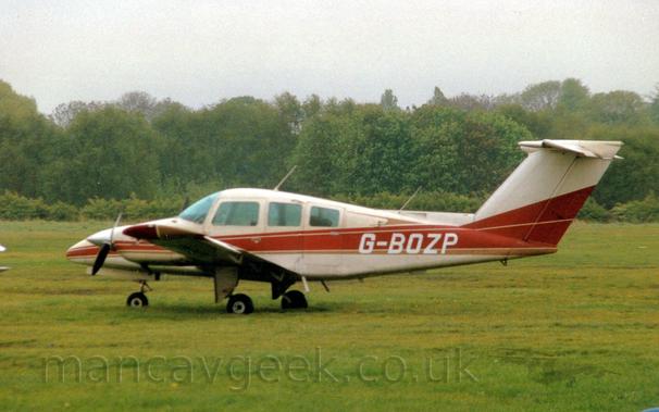 Side view of a cream-coloured, twin engined light aircraft parked on grass facing to the left.
There is a brown stripe running along the body, getting wider as it moves to the rear, overlaid with the white registration "G-BOZP" on the rear fuselage.
Green grass fills most of the frame, with rows of trees in the distance, under grim grey skies.