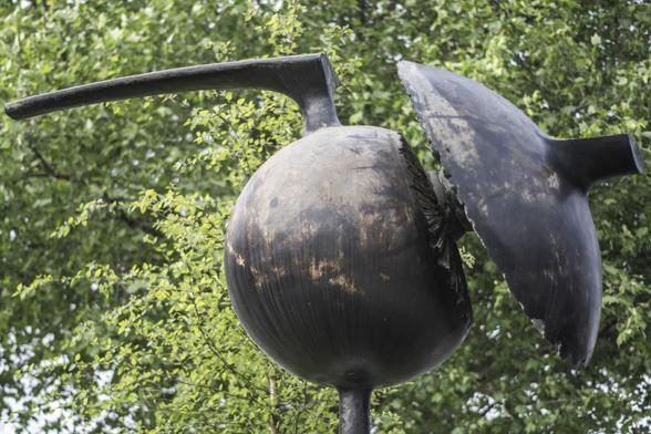 Photo of a sculpture in metal (bronze I think?) in front of a green tree. The sculpture is a round ball with a part off to the side that looks like the top of an acorn, and a kind of handle or beak on the top. Abstract.