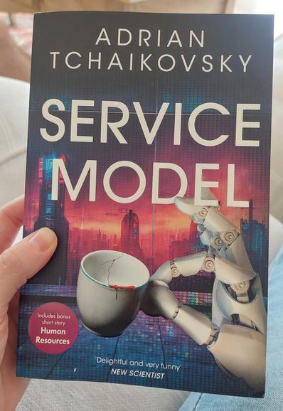 A photo of my hand holding the book Service Model by Adrian Tchaikovsky. The cover is of a robot hand delicately holding a teacup but the teacup has a mysterious red liquid dripping down the edge.