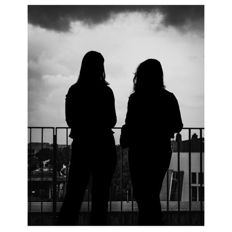 Zwei Silhouetten weiblicher Figuren stehen nebeneinander an einem Geländer. Sie blicken in die Ferne, während ein bewölkter Himmel über ihnen hängt. Im Hintergrund sind anonyme städtische Gebäude und Bäume sichtbar, die in die Dämmerung getaucht sind. Die Szene strahlt eine ruhige, nachdenkliche Atmosphäre aus und lässt Raum für Interpretationen über die Gedanken der Figuren.