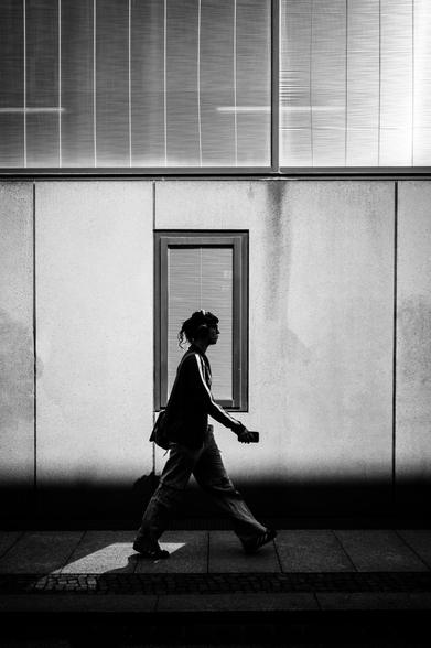 The image captures a lone figure walking along a sidewalk adjacent to a building in a city environment. The person is silhouetted against the light, making details difficult to discern. In the background, a large window with closed blinds looms, adding a sense of uniformity and enclosure to the scene. The foreground consists of the sidewalk and the person's shadow, stretching out before them.