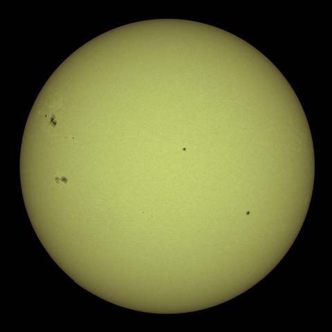 A yellowish image of the Sun with several visible sunspots scattered across its surface, set against a black background.