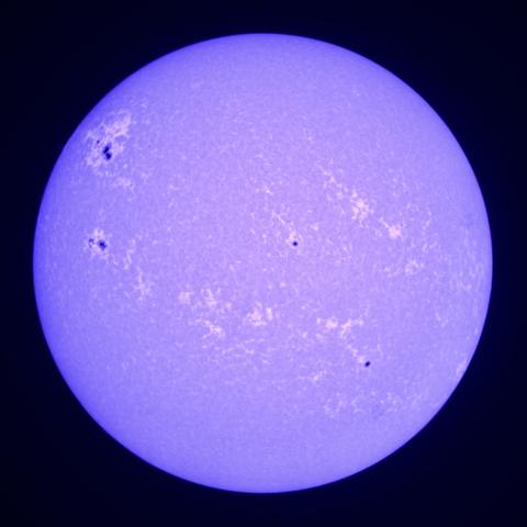 A purple-tinted image of the Sun showing several dark sunspots and surface texture against a black background.