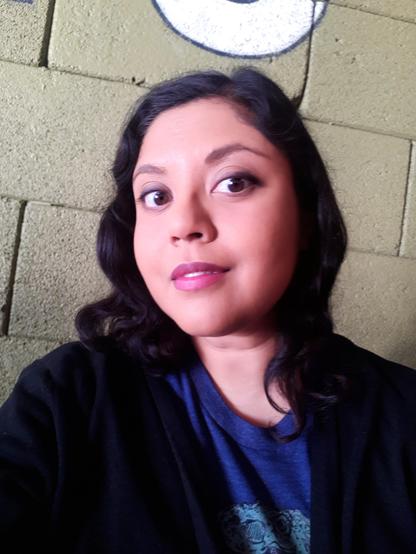 Head-and-shoulders color photo of author Premee Mohamed, a woman of Indo-Caribbean ancestry. She wears her black hair without bangs, at shoulder length, in glossy waves. She wears a black cardigan over a blue tee, smoky cat-eye makeup, and a dark pink lipstick. She stands in front of a gray cinderblock wall. She looks down and to the side into the camera, her eyebrows arched and her large brown eyes wide, smiling slightly.
