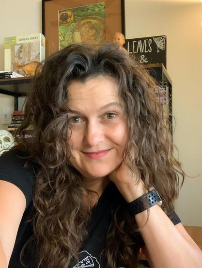 Head-and-shoulders color candid photo of author Victoria Nations, a fair-skinned woman with light hazel (?) eyes and curly brown hair worn long without bangs, the better to show off her widow’s peak. She wears a black tee and a watch with a wide black band. She sits in front of a bookshelf that includes books, an Edward Gorey jigsaw puzzle, and a creepy baby doll. She leans on one hand and quirks an eyebrow with her smile into the camera.