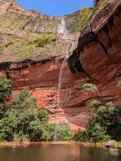 Der Betrachter steht unterhalb einer steilen rötlichen Felswand. Ein Wasserfall mit wenige Wasser stürzt aus großer Höhe in ein Becken. Um das Becken und an den Felswänden wachsen Bäume und Sträucher. Blauer Himmel oberhalb der Felsen.