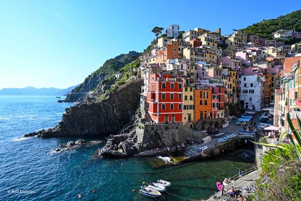 A picturesque coastal village with colorful buildings nestled on hillside cliffs, overlooking a sparkling blue sea. Small boats and people are along the rocky shoreline, with lush green hills in the background under a bright blue sky.