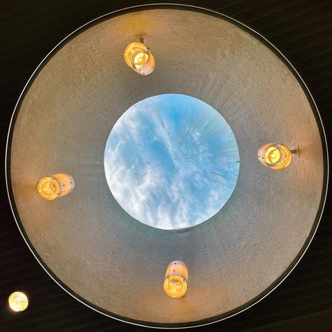 View from below a circular skylight, showing blue sky and clouds, surrounded by four decorative lights.
