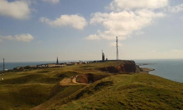 Grünfläche oben auf helgoland, am Rand rote Felsen, im Hintergrund Leuchtturm, Funkmast und Ort mit Kirchturm