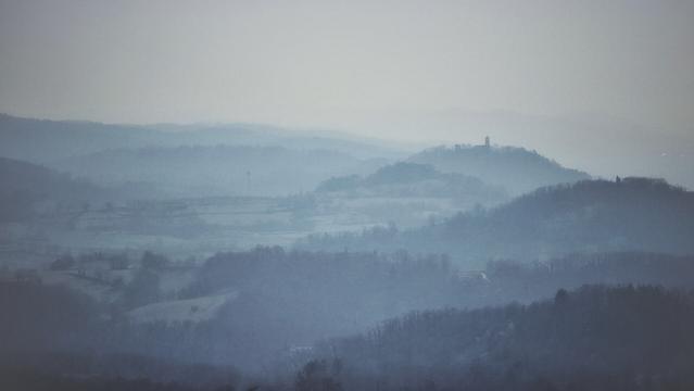The image is a landscape photograph featuring multiple layers of rolling hills extending into the distance under a hazy sky. The color palette is predominantly muted blues and grays, contributing to an overall cool tone. The foreground shows silhouetted tree lines, with some indistinct shapes that could be structures or open fields. As the hills recede, they become progressively lighter and less distinct due to atmospheric perspective and mist. On one of the more distant hilltops, a tower is visible. The lighting is soft and diffuse, due to an overcast day.