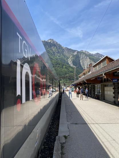 A train at a platform. The TGV inOui branding is prominent on the carriage on the left, a mountain below a blue sky in the background and train station buildings on the right