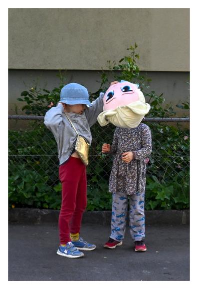The image depicts two children outdoors, standing near a fence with greenery in the background. The child on the left is wearing a light gray long-sleeve shirt, red pants, blue shoes, and a blue bucket hat. They are also carrying a small gold crossbody bag. The child on the right is dressed in a dark floral-patterned dress over gray pants with blue patterns, and red shoes. The child on the left holds a large puppet head with blonde hair, covering the head of the other child. The scene appears to be playful and casual.