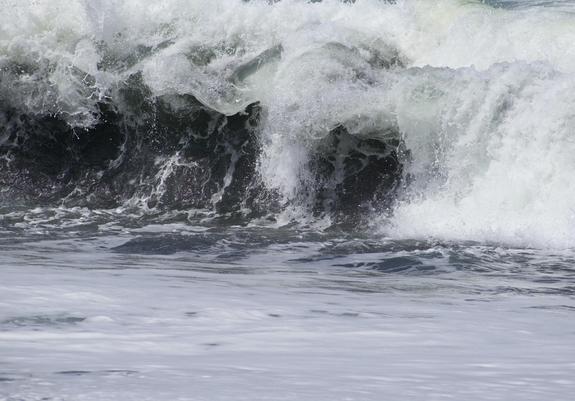 Close up shot of a wave crashing, surrounded by white water. Rounded roiling white water behind the wave, and flat white foaming water in front of it, bracketing the small violently crashing curl of wave. 

On one side the curl is closing, creating a rounded white roil of water. On the other side, the wave is still cresting and curling over, green and white curling shapes tipping over the top and starting to form droplets and puffs and splatters of water overhanging the wave beneath. The wave must be pulling up sand, because the wave under the curl is very dark at a few points, dark greenish browns in the shadow of the curling water above them. They almost look like cave entrances under the wave’s curling top.