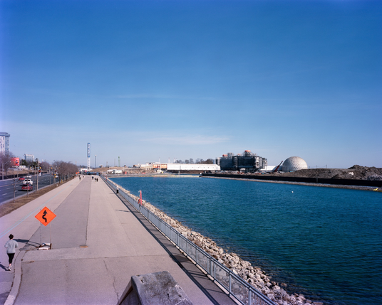 At the lower left, a runner passes a hazard zigzag sign. To their left, the broad lanes of Lake Shore Boulevard, to their right, the west channel, and, before them, the destruction of the landmarks of Ontario Place.