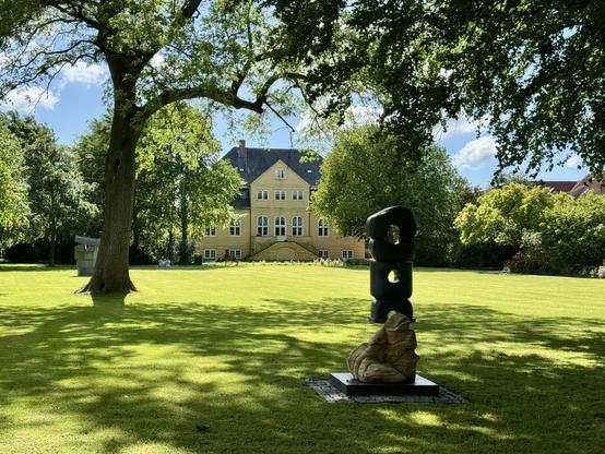 A sunny outdoor scene featuring a large green lawn and a yellow building in the background. In the foreground, there are two sculptures—one made of dark material and another of light-colored stone—surrounded by trees and manicured greenery.