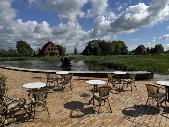 A scenic outdoor area with several empty tables and chairs on a brick patio, overlooking a pond with greenery and a decorative fountain in the shape of an opening flower. In the background, there are two houses and a clear blue sky with scattered clouds.