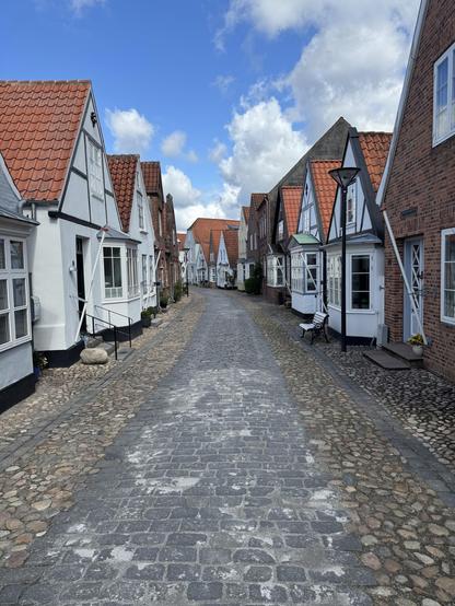 A picturesque, narrow cobblestone street lined with charming, traditional houses featuring sloped roofs and a mix of white and brick facades. Bright blue skies with scattered clouds create a serene atmosphere.