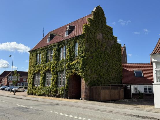 A large ivy-covered building with a red tiled roof stands beside a street. It has multiple windows and a weather vane on top. In the background, there are additional buildings and some parked cars under a blue sky with clouds.