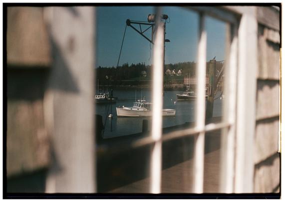 A film photo of a window on a wooden-shingled building on a wharf. The reflection in the window is the primary subject of the photo. A harbor scene is reflected with three or four boats on moorings visible and an old sardine cannery building in the background.
