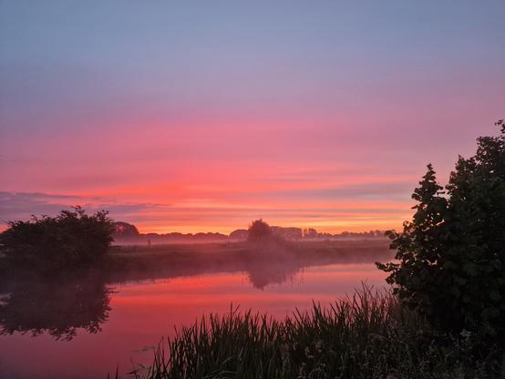 AI Alt Text:

A serene early morning landscape captures the vibrant colors of a sunrise reflecting on a still, mist-covered body of water. The sky is painted in vivid layers of purples, pinks, oranges, and hints of yellow, gradually transitioning into a soft blue at the top. Gentle morning fog hovers over the fields in the distance, adding an ethereal atmosphere. Silhouettes of trees and bushes frame the scene on both the left and right sides, while tall grasses and reeds in the foreground are lightly dusted with dew. The reflection in the water mirrors the colorful sky, enhancing the tranquil and dreamlike mood of the image.