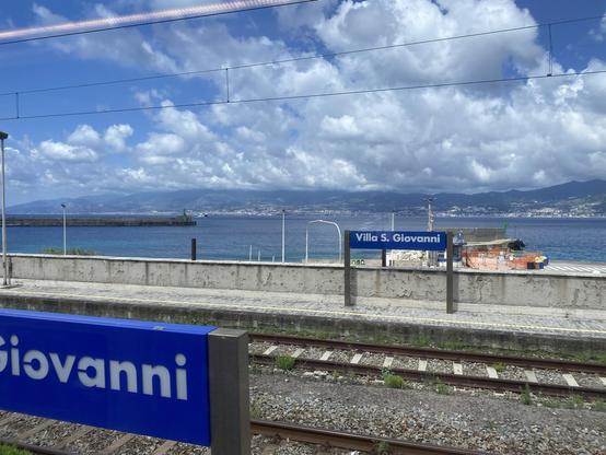 Station boards reading Villa S. Giovanni in a train station with a spectacular view across the Straits of Messina. The sea is immediately behind an open train platform. A harbour wall extends from the left as a distant ferry approaches the harbour. Across the strait the shore is lined with buildings backed by steeply rising hills. The sky has large masses of bright white cloud and patches of bright blue.