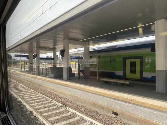 A platform at Milano Porta Garibaldi. Empty rail tracks in the foreground with a covered platform and green and blue train at the other side of the platform. Station signs in white lettering on a blue background show the station name