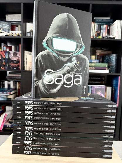 Les 11 tomes de Saga empilés devant une bibliothèque et le 12eme posé dessus. En couverture, un personnage à tête d’écran dans un hoodie à capuche noir qui regarde sa « main » en forme d’arme.