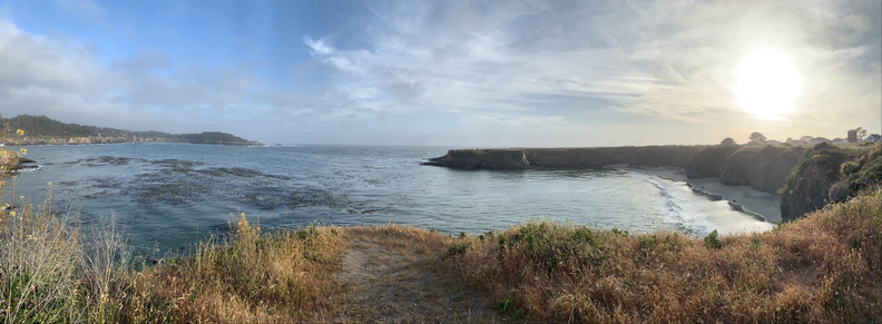 A view of the Pacific coastline near Mendocino, California. The viewer is on a grassy cliff overlooking a small bay formed by a peninsula reaching out from the right. The sky is blue with wispy scattered clouds and the sun approaching the horizon but not yet setting.