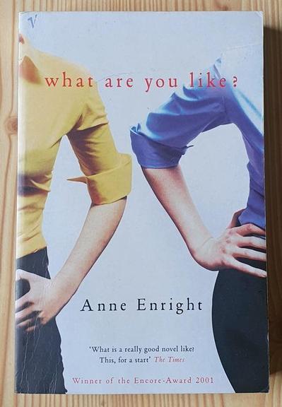 Paperback copy of Anne Enright's novel "What Are You Like?" It shows two young women side by side, hands on hips, but we can only see their shoulders, arms, and hips. One wears a yellow shirt, the other, blue, both with the sleeve rolled up. A cover quote from the Times reads: "What is a really good novel like? This, for a start." Below that is the text: "Winner of the Encore Award 2001". The photo is by Ashley Rudolph.