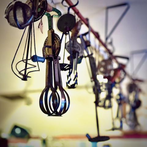 Multiple older whisks and other kitchen utensils hang in the dim light against a bright wall in the distance.