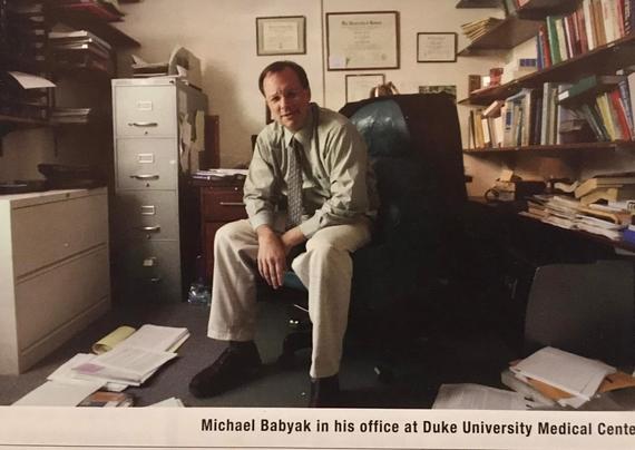 Man in an office chair surrounded by scattered stacks of books and papers.
