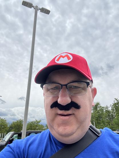It’sa me! Wearing a red cap with an "M" logo and a fake mustache, dressed in a blue shirt, poses in an outdoor setting with overcast skies and vehicles in the background.