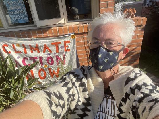 Masked woman wearing a Climate Emergency tshirt stands in front of a homemade Climate Action Now sign