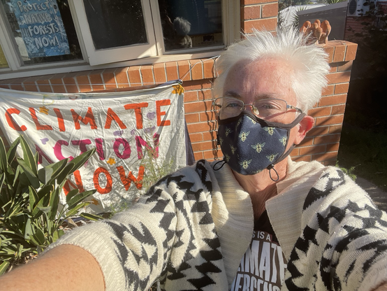 Masked woman wearing a Climate Emergency tshirt stands in front of a homemade Climate Action Now sign