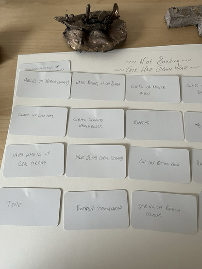 Image shows multiple small white cards on paper, each with a description written on. For example “Cup on beach rock” or “waves washing up coral strand”. In the background is a bronze sculpture of a ship.
