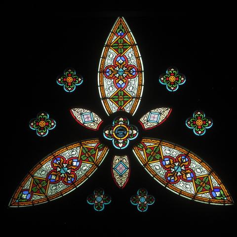 A colourful stained glass window. Only the window is visible, the rest of the photgraph is black.
The main colours are white, magenta, red, green and orange. The design has three large lobes and three smaller lobes. Each lobe has floral and vegetal decorations as well as geometric shapes likes squares. There are also six four-lobed shapes between the large lobes: a similar but larger motif is in the centre. It has the text “Rousseau & Allard. France. Angers 1908.”