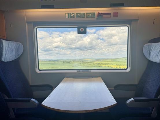 Picture of the interior of the second class cabin of an ICE-T, showing four empty chairs round a table. Outside the window an expansive green landscape can be seen, topped by a cloudy sky