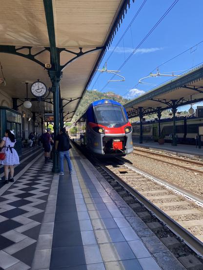 A blue, red and grey liveried modern train arriving at a train platform which is paved in black and white patterned tiling. The platform canopy is in an old ornate style with white wooden panels supported by dark painted iron columns. Passengers prepare to board while a clock fixed to one of the columns reads 2:14pm