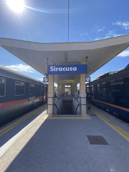 A station platform with the station name, Siracusa, in white lettering on a blue background above a stairway leading downwards. Above this the thick white concrete? platform canopy stretches out and gently upwards. Above a bright white sun shines from a mostly blue sky. A train is stopped at each side of the platform, the Intercity Notte on the left and the Regionale Veloce on the right.