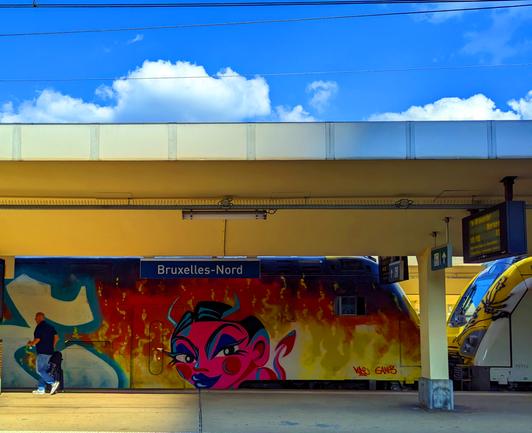 A person is walking on the platform of Bruxelles-North station; a train with graffiti in colorful motifs stands under a bright blue, cloudy sky.

Eine Person geht auf dem Bahnsteig des Bahnhofs Bruxelles-Nord; ein Zug mit Graffiti in bunten Motiven steht unter einem strahlend blauen, bewoelkten Himmel.