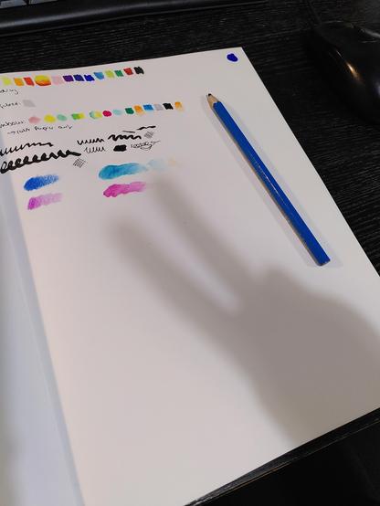 A sketchbook page where I tested various pens and pencils. There is a blue pencil lying on the page. also there is the shadow of my hand forming a ✌️