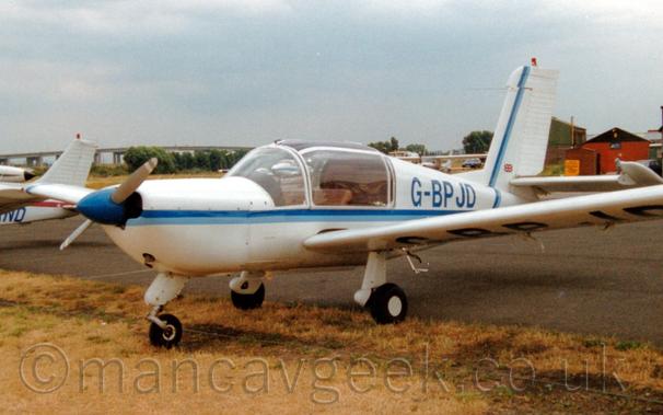 Side view of a single-engined light aircraft parked facing to the left on yellowing grass, right on the edge of a black tarmac taxiway.
The plane is mostly white, with a 2-tone blue stripe running along the body before sweeping up the front of the tail.
The registration "G-BPJD" is on the upper rear fuselage, as well as under the left wing.
Another couple of planes are parked around it, as well as a couple of wooden buildings on the right.
A long bridge reaches over the horizon on the left, with blue-grey sky filling the rest of the frame.