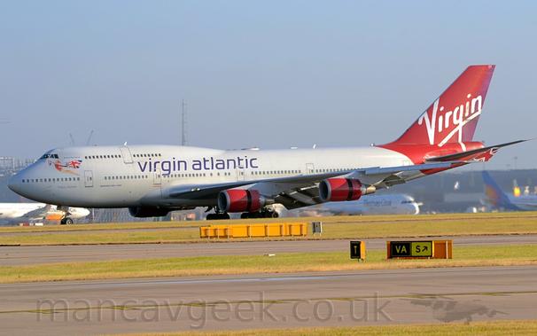 Side view of a very large, white, 4 engined jet airliner moving from right to left .
The plane is mostly white, with purple "Virgin Atlantic" titles on the upper forward fuselage, and a scarlet red rear fuselage and tail, with white "Virgin" titles.
The upturned wing-tips have part of a waving Union Flag.
The engine pods slung under the wings are the same scarlet red, with the rear 1/3rd of the cowling having moved backwards to expose the thrust reversers, which along with the flaps hanging from the rear of the wings is slowing the plane after landing.
Taxiways lined with strips of grass fill the foreground, with a grey terminal building with several planes parked in front of it stretching across the frame, under grey-blue skies.